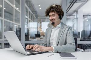 lächelnd jung Geschäftsmann mit Headset Arbeiten auf Laptop im modern Büro, präsentieren Multitasking und Kunde Bedienung Fähigkeiten. positiv Arbeit Umgebung reflektiert im seine heiter Ausdruck. foto