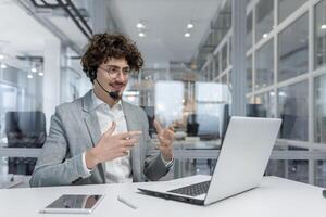 lockiges Haar Geschäftsmann unterhält auf Headset während Arbeiten auf Laptop im ein modern Büro Einstellung, zeigen Professionalität und Sachverstand. foto