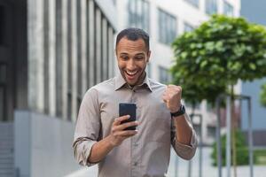 afrikanisch amerikanisch Geschäftsmann draußen Büro Gebäude mit Telefon, lächelnd und glücklich halten Hand oben Triumph Geste, jung Unternehmer feiern Sieg lesen gut Investition Nachrichten online foto