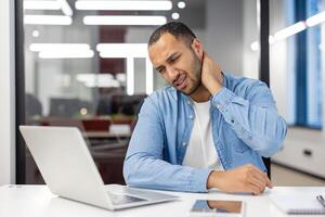 müde spanisch Mann im Hemd Sitzung im Büro beim Schreibtisch mit Laptop und halten Hand auf Nacken, tun Massage, Gefühl Druck und Schmerz. foto