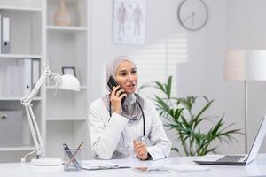 zuversichtlich Muslim weiblich Arzt im Hijab Sprechen auf das Telefon, Sitzung im ein gut beleuchtet modern Klinik Büro. sie sieht aus Fachmann und zugänglich. foto