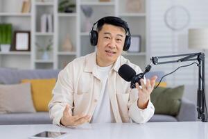 freundlich asiatisch Mann mit Kopfhörer Podcasting, reden und gestikulieren gegenüber das Kamera mit ein Übertragung Mikrofon und ein Laptop im ein Zuhause Studio Einstellung. foto