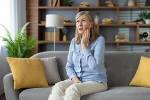 kämpfen reifen Dame Gefühl Dental Schmerzen und Reiben Wange mit Palme während Sitzung mit öffnen Mund. blond weiblich haben Zahn schmerzen und erfordern Besuch zu Klinik zum bekommen loswerden von Entzündung. foto