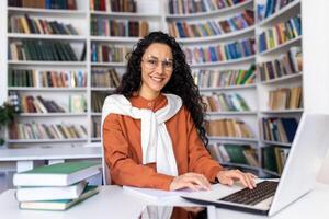 Porträt von heiter lächelnd Mädchen Student, Frau suchen beim Kamera Sitzung studieren mit Laptop, spanisch Frau tragen Brille studieren online im Universität Bibliothek unter Brille mit Laptop. foto