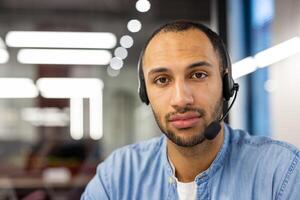 Porträt von ein jung arabisch Mann Sitzung im ein Headset im Vorderseite von ein Laptop im das Büro und selbstbewusst suchen beim das Kamera. Nahansicht Foto. foto