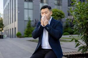 krank jung asiatisch Mann im ein passen Sitzung auf ein Bank in der Nähe von das Büro und abwischen seine Nase mit ein Gewebe von ein flüssig Nase, Leiden von saisonal Allergien. foto