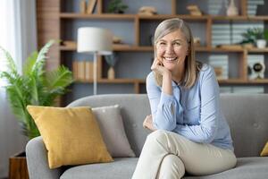 heiter reifen Frau Sitzung auf dekoriert Sofa mit gekreuzt Beine und halten Kinn mit Palme. positiv weiblich im beiläufig Kleider suchen beim Kamera während Ausgaben Tag beim gemütlich Wohnung Innere. foto