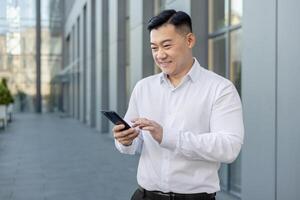 asiatisch lächelnd jung männlich Büro Arbeiter Stehen draußen im Weiß Hemd und mit Handy, Mobiltelefon Telefon. foto