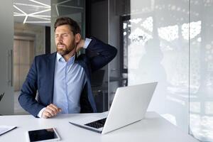 müde jung Geschäftsmann Arbeiten im das Büro mit ein Laptop, Sitzung beim das Tabelle und halten seine Nacken, verzog das Gesicht im Schmerz, tun ein Massage. foto