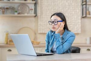 müde jung asiatisch Frau Freiberufler Sitzung beim ein Tabelle mit ein Laptop, gelangweilt, unfähig zu Start arbeiten. funktioniert entfernt beim Zuhause während Quarantäne. foto