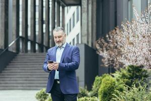 Senior grau haarig Mann, ein Geschäftsmann im ein Anzug, steht in der Nähe von das modern Büro Center und Anrufe ein Taxi von das Telefon, Tippen das Nummer, wartet, ist im ein Eile zu treffen. foto