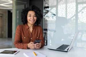 Porträt von erfolgreich Geschäftsfrau beim Arbeitsplatz, weiblich Boss lächelnd und suchen beim Kamera, reifen und zufrieden Latein Frau Arbeiten Innerhalb Büro mit Laptop beim Arbeit halten Telefon im Hände. foto