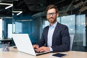 Porträt von reifen Boss Innerhalb Büro mit Laptop, erfolgreich und zufrieden Investor Manager suchen beim Kamera und lächelnd Mann im Brille und Geschäft Anzug, Investor mit Bart Sitzung auf Stuhl foto