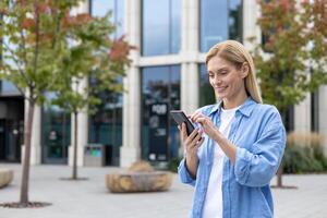 reifen Frau mit Telefon im Hände Gehen im das Stadt, ein Geschäftsfrau im Blau Hemd halten Smartphone im Hände, lesen online Sozial Netzwerke, blond lächelnd Befriedigung ist mit ein Anwendung. foto