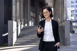 zuversichtlich jung erfolgreich Geschäft Frau Stehen draußen ein Büro Center und Gerichtsgebäude, halten ihr Hand im ihr passen Tasche und suchen lächelnd beim das Handy, Mobiltelefon Telefon Bildschirm. foto