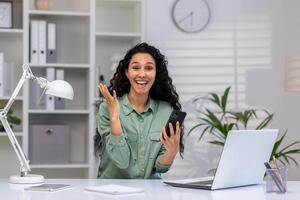 begeistert reifen spanisch Dame Multitasking im ein hell Zuhause Büro, Arbeiten auf Laptop und chatten auf Telefon mit ein lächeln. foto