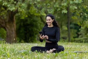 lächelnd Frau gekleidet im schwarz Activewear Sitzung auf Gras, einnehmend mit ihr Smartphone im ein üppig Grün Park. foto
