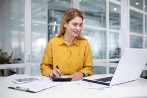 erfolgreich reifen weiblich finanziell Buchhalter beim Arbeitsplatz Innerhalb Büro, füllt Daten im Tisch, funktioniert mit Unterlagen und Papiere, Verwendet Laptop und Taschenrechner zum Papierkram. foto