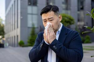 Nahansicht Foto von ein krank jung asiatisch männlich Geschäftsmann Sitzung auf ein Bank in der Nähe von ein Büro Gebäude und abwischen seine Nase, mit ein Serviette von ein flüssig Nase.