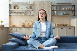 schön asiatisch Frau entspannend beim Zuhause Sitzung auf Sofa und meditieren im Lotus Pose, tun Atmung Übungen im Leben Zimmer. foto