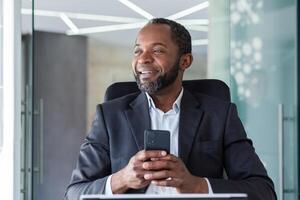 Nahansicht Foto von ein lächelnd Afroamerikaner männlich Geschäftsmann Sitzung beim ein Büro Schreibtisch im ein Geschäft passen und mit ein Handy, Mobiltelefon Telefon, suchen zu das Seite.