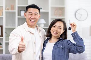 glücklich jung asiatisch Paar gekauft ein Wohnung, ein Haus. Sie Stand umarmen und lächelnd beim das Kamera. das Mann zeigt an Super mit seine Finger, das Frau hält das Schlüssel im ihr Hand. foto