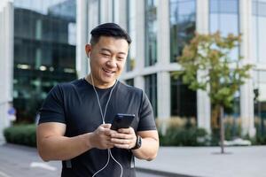 ein jung Mann, ein asiatisch Sportler, ist Stehen auf ein Stadt Straße im Kopfhörer, halten ein Telefon im seine Hände, Herstellung ein Forderung. Zifferblätter Mitteilungen, chattet. foto