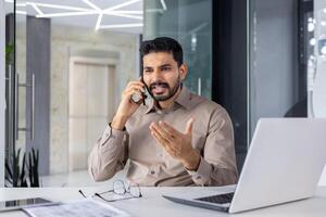 ein jung männlich Fachmann erscheint wütend während ein Telefon Anruf im ein modern Büro Einstellung. er Gesten mit seine Hand, zeigen Frustration. foto