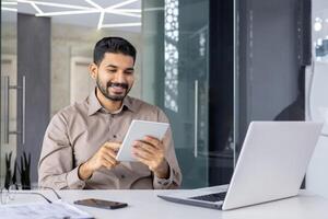 lächelnd Geschäftsmann im ein Beige Hemd mit ein Tablette während Sitzung beim ein Schreibtisch geschmückt mit ein Laptop, Smartphone, und Unterlagen im ein stilvoll Büro. foto
