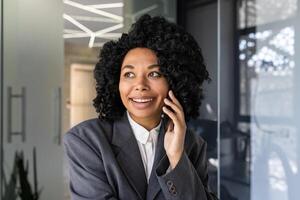 Nahansicht Porträt von ein jung schön Afroamerikaner Frau Sitzung im das Büro im ein Geschäft passen und lächelnd reden auf das Telefon. foto