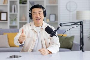 heiter asiatisch Mann im ein Headset mit Mikrofon geben ein Daumen oben während suchen beim das Kamera, Anzeige ein positiv Konversation oder erfolgreich Podcast Aufzeichnung. foto
