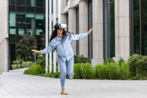 jung schön Mädchen im Hemd und Jeans Tanzen draußen Büro Gebäude tragen virtuell Wirklichkeit Gläser, spanisch Frau glücklich haben Spaß und entspannend mit lockig Haar. foto