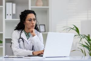 ein Fachmann spanisch weiblich Arzt ist aufmerksam Arbeiten auf ihr Laptop im ein modern Klinik Büro, Anzeigen Widmung und Pflege. foto