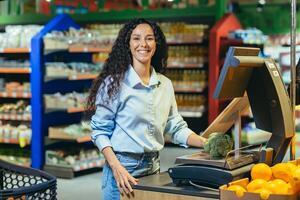 Porträt von ein glücklich Frau Käufer im ein Supermarkt, ein spanisch Frau wiegt Brokkoli Kohl auf ein Skala, lächelt und sieht aus beim das Kamera, im ein Geschäft unter Regale mit Lebensmittelgeschäft Produkte foto