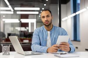 Porträt von ein jung ernst Muslim Mann Sitzung im das Büro beim ein Schreibtisch mit ein Notizbuch und Unterlagen, halten ein Tablette im seine Hände und suchen Ernsthaft in das Kamera. foto