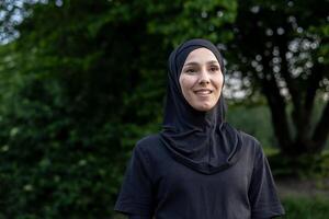 Porträt von ein lächelnd Muslim Frau tragen ein Hijab draußen, ausströmend Vertrauen und Gelassenheit im ein still Park Rahmen mit Grün Laub Hintergrund. foto