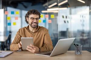 lächelnd männlich Fachmann Multitasking mit ein Tablette und Laptop im ein modern Büro Rahmen mit klebrig Anmerkungen im das Hintergrund. foto