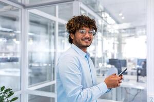 Porträt von erfolgreich Geschäftsmann Innerhalb modern hell Büro, indisch Mann lächelnd und suchen beim Kamera, Mann im Hemd und Brille halten Telefon. foto