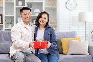 ein jung asiatisch Paar, ein Mann und ein Frau, sind Sitzung auf das Sofa beim Zuhause und halten ein Geschenk Box im ihr Hände. lächelnd und glücklich suchen beim das Kamera. foto