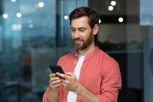 lächelnd und glücklich Geschäftsmann Innerhalb das Büro durch das Fenster Verwendet das Telefon ein reifen Mann mit ein Bart sieht aus beim das Smartphone Bildschirm, durchsucht Internet Seiten, Typen Mitteilungen im ein online Anwendung foto