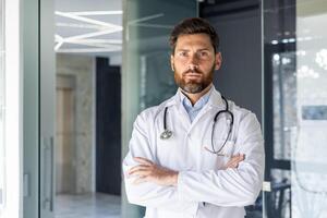 Porträt von ein selbstbewusst männlich Arzt Stehen im ein Krankenhaus Zimmer im ein Weiß Mantel, Kreuzung seine Waffen auf seine Truhe und Ernsthaft suchen beim das Kamera. foto