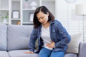 asiatisch Frau allein beim Zuhause krank Sitzung auf Sofa, hat stark Bauch Schmerz, nicht wohl Frau hält ihr Hand durch ihr Seite im das Leben Zimmer von das Haus. foto