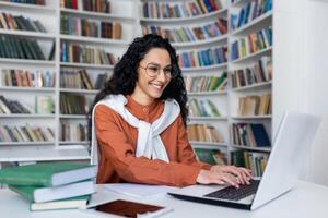 lächelnd beiläufig Brünette Tippen auf Laptop Tastatur während Sitzung im Campus Bibliothek mit akademisch Bücher beiseite. vielversprechend Absolvent Schüler Fertigstellung Artikel und Gefühl zufrieden mit Forschung Ergebnis. foto