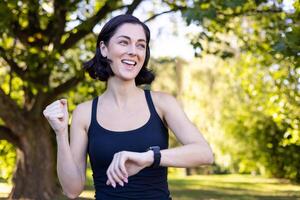 Nahansicht Foto von glücklich jung Frau tun Sport und Joggen im Park, suchen beim Clever Uhr und zeigen Erfolg Geste mit Hand, Jubel im Sieg.