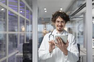 ein Arzt im ein Weiß medizinisch Mantel und Stethoskop hält ein Tablette zum ein medizinisch konsultieren im seine Büro. Lächeln Mann suchen beim Kamera foto