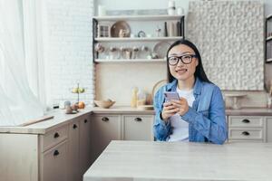 ein glücklich jung asiatisch Teenager Mädchen ist Sitzung beim Zuhause im das Küche, halten ein Telefon im ihr Hände und Senden ein Botschaft zu ihr geliebt eins, ein Freund. suchen beim das Kamera, lächelnd. foto