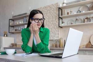 ein jung verärgert Frau sitzt beim Zuhause im das Küche mit ein Laptop, weint, Tücher ihr Tränen mit ein Serviette foto