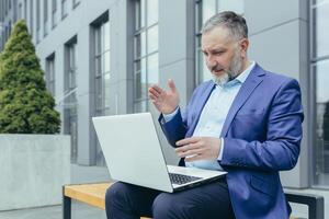 erfolgreich Senior männlich Geschäftsmann führt ein Konferenz von ein Laptop. Sitzung draußen im ein passen auf ein Bank in der Nähe von ein Büro Center, Wellen seine Hände, erklärt. foto