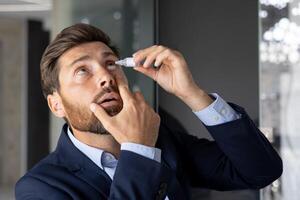 Nahansicht Foto von ein jung Geschäftsmann Sitzung im das Büro beim ein Schreibtisch im ein passen und einflößen Medizin in seine Auge, Behandlung Entzündung, Schmerz, chronisch Ermüdung.