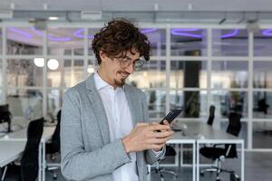 ein jung Mann funktioniert im ein Büro, steht Innerhalb und Verwendet ein Handy, Mobiltelefon Telefon mit ein lächeln, Tippen Mitteilungen, SMS schreiben. foto